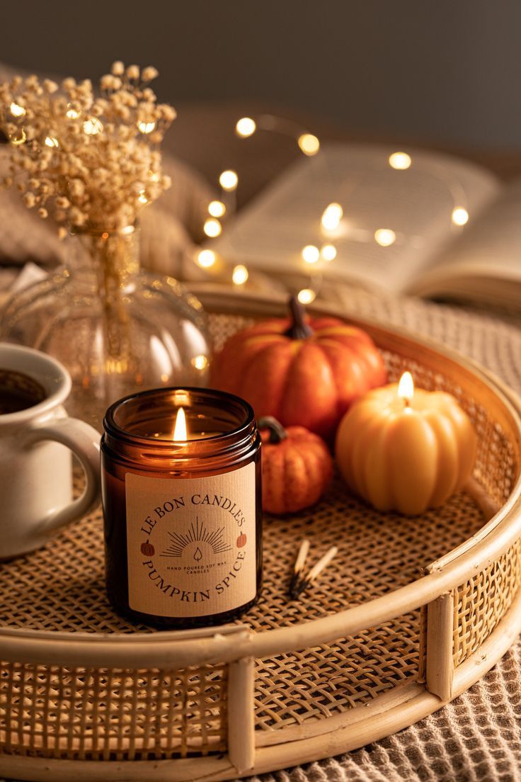 Rustic scandi wooden tray table with mini pumpkins, scented candles, cup of hot drink, and pampas grass on it to mimic cosy, dark evenings of colder seasons. Paired beside it lays a half open book.